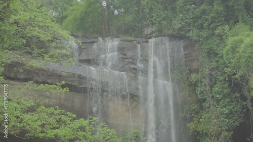 A ungraded zoomed medium to close shot scenery shot at daylight of a big rocky waterfall with running water surrounded by green trees and moss in a forest or jungle.
