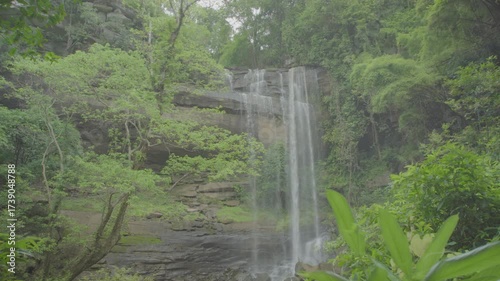 A ungraded wide establishment scenery shot at daylight of a big rocky waterfall with running water surrounded by green trees and moss in a forest or jungle.