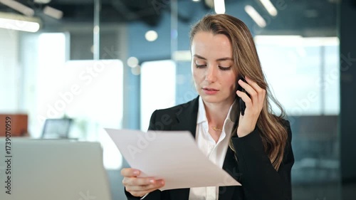 Focused businesswoman in a black blazer and white shirt makes a serious phone call while examining documents in a modern office. She appears focused and busy.
