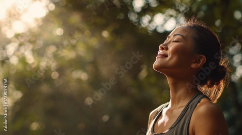 Medium view of a healthy Asian adult outdoors, standing in nature, closing eyes while taking a deep breath of fresh air, smiling with a calm and energized expression, posture open and relaxed
