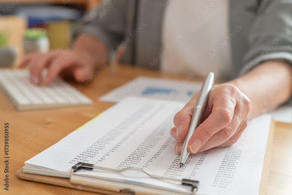 Naklejka premium Person using a pen to review financial numbers on a clipboard, while simultaneously operating a computer keyboard, focusing on data analysis and strategic planning