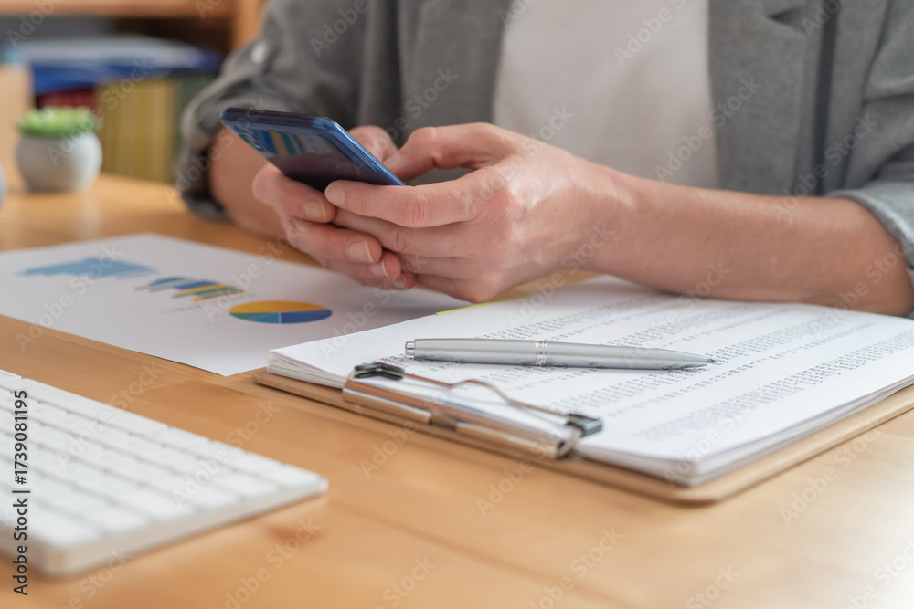 Fototapeta premium Businesswoman's hands holding a smartphone while checking financial charts and reports on documents on a wooden desk, representing modern data analysis and business communication