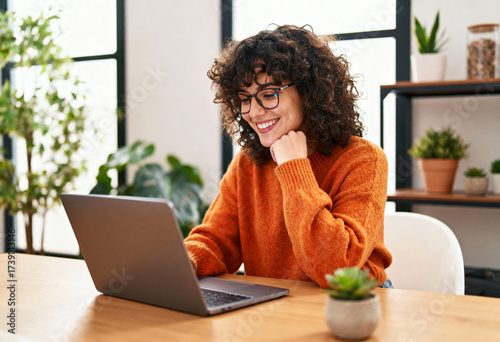 A woman in an orange sweater smiles while using a laptop in a well-lit room with plants.