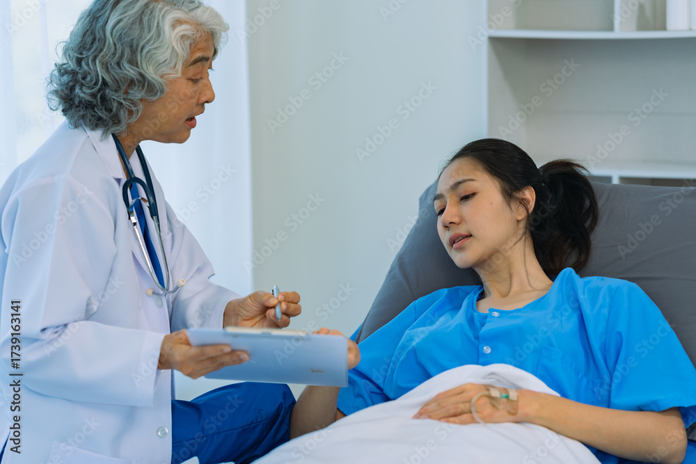 Obraz premium Elderly female doctor and Asian female patient are discussing something while sitting on hospital bed. Medical consultation concept.