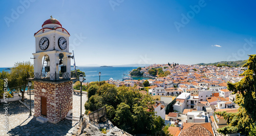 Iconic clock tower with red dome and stone base overlooking Skiathos Town and the Aegean Sea. Yachts and boats are visible in the harbor, surrounded by traditional white houses with terracotta roofs.
