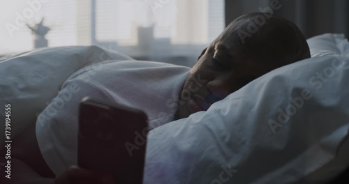 Close up of Man in Bed, Resting Head on White Pillow, Scrolling Through Social Media Content in Bright Sunlight. Concept of Connected Lifestyle, Morning Digital Routine, Digital Habits. Static Shot.