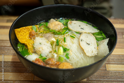 Close-up of a delicious Thai noodle soup served in a black bowl. The dish includes rice noodles, fish balls, sliced fish cakes, crispy wonton, fresh herbs, and clear flavorful broth. Authentic Asian s