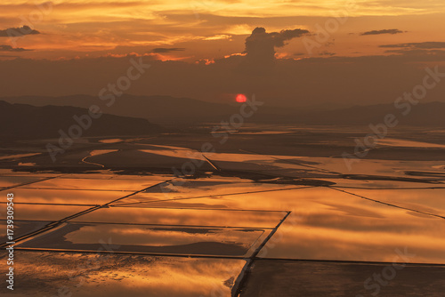 Aerial view of the setting sun casting a fiery glow over the geometric salt ponds, painting the landscape in shades of orange and gold, Salt Lake City, Utah, United States.