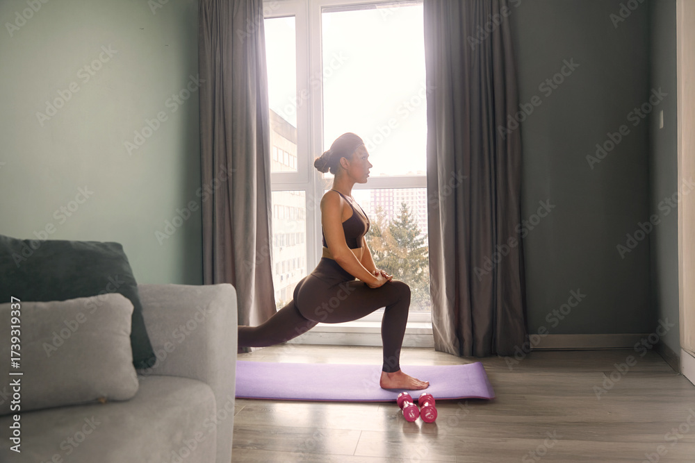 Fototapeta premium Woman practicing lunges on a yoga mat during morning workout in bright room with large window