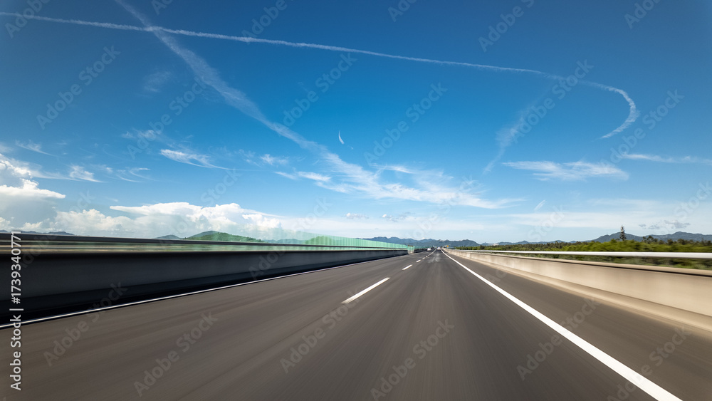 Naklejka premium Highway stretches under clear blue sky with clouds and distant mountains