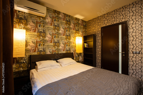 Apartment interior. Double bed in the bedroom. Pillows and bedspread on the made bed. Wallpaper with images of European landmarks. Air conditioning and wall lamps on the wall. Door in the wall.