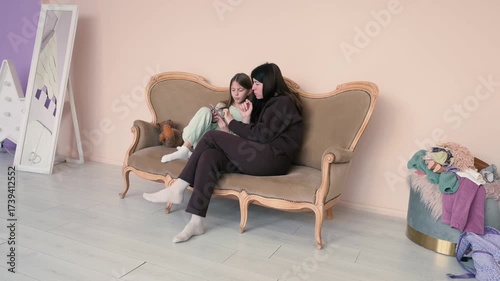 Mother and daughter share a tender moment on a vintage sofa, engaging with a smartphone. This candid scene captures family bonding and modern digital connection in a cozy home setting.