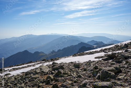 Snow-dusted rocks and layered mountain ridges in Sierra de Gredos, Spain, under a soft blue sky. The expansive view highlights the untouched wild beauty and tranquil atmosphere.