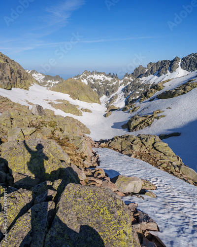 Rocky ridge and snowfields in Sierra de Gredos, Spain, with mountain peaks under a vivid sky. A shadow of a hiker appears on the rocks, capturing the wilderness adventure.