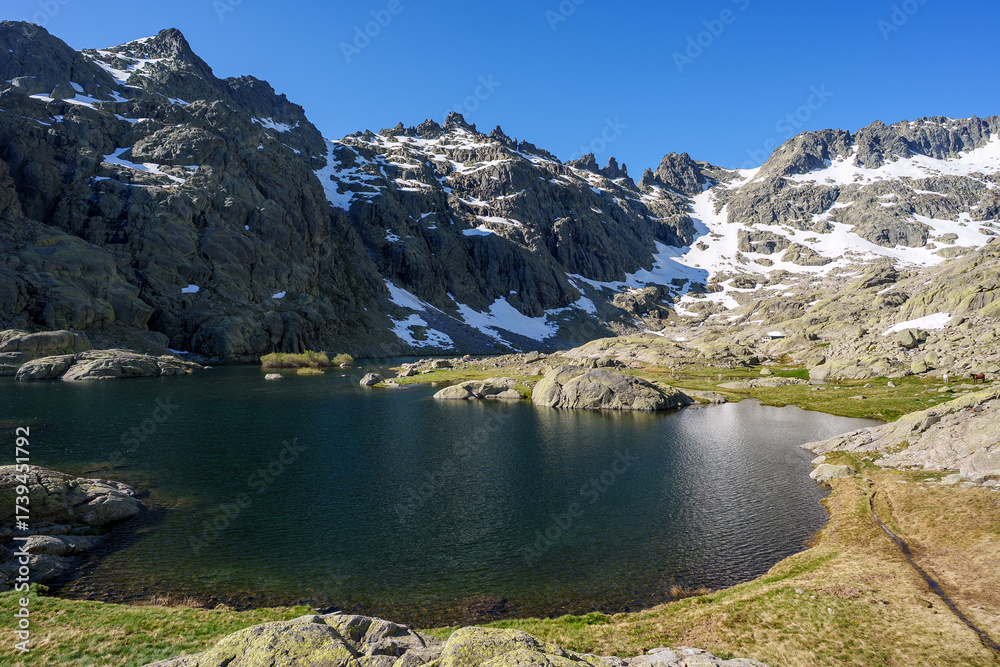 Obraz premium Laguna Grande in Sierra de Gredos, Spain, with deep blue water, rocky slopes, grassy shore, and snow patches on jagged mountain peaks under clear blue sky in spring.