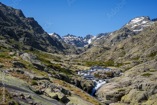 Rocky hiking path in Sierra de Gredos, Spain, with snow-capped peaks, clear blue sky, mountain stream, and dramatic rugged landscape perfect for adventure and nature lovers.