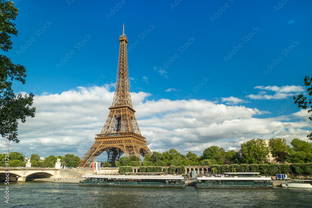 Fototapeta premium Eiffel Tower in Paris with Seine River and blue sky, symbolizing romance, architecture, travel, tourism, European landmark, and cultural heritage under scenic urban atmosphere.
