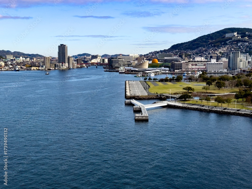 Obraz premium Der Hafen von Nagasaki im Sonnenlicht mit ruhigem Wasser, kleinen Booten und Blick auf die umliegenden Stadtteile. Ein idyllischer Ort, der das maritime Erbe Nagasakis widerspiegelt