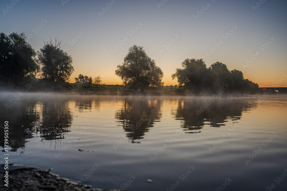 Fototapeta premium Stimmungsvolle Morgenaufnahme am Fluss mit nebligem Wasser und Bäumen im Licht der Morgendämmerung