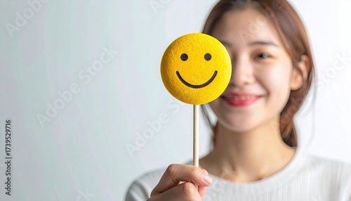 Smiling Young Woman Holding a Yellow Smiley Face on a Stick in Front of a White Background with Soft Lighting Happy Expression and Blurred Figure