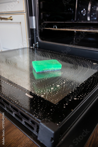 Oven door is lowered down and washed. Sponge for washing lies on its surface