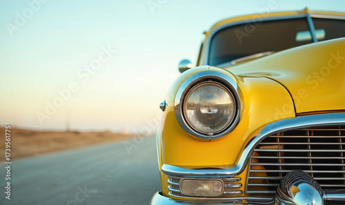 Close up of a bright yellow vintage car headlight and chrome grille parked on an empty road during sunset. The classic automobile gleams with a retro style.