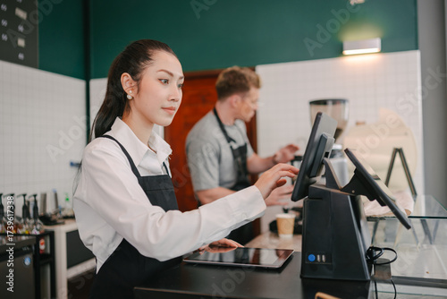 A dedicated barista focuses on the point-of-sale tablet while a coworker prepares a drink in the background, showing the teamwork involved in a coffee shop.