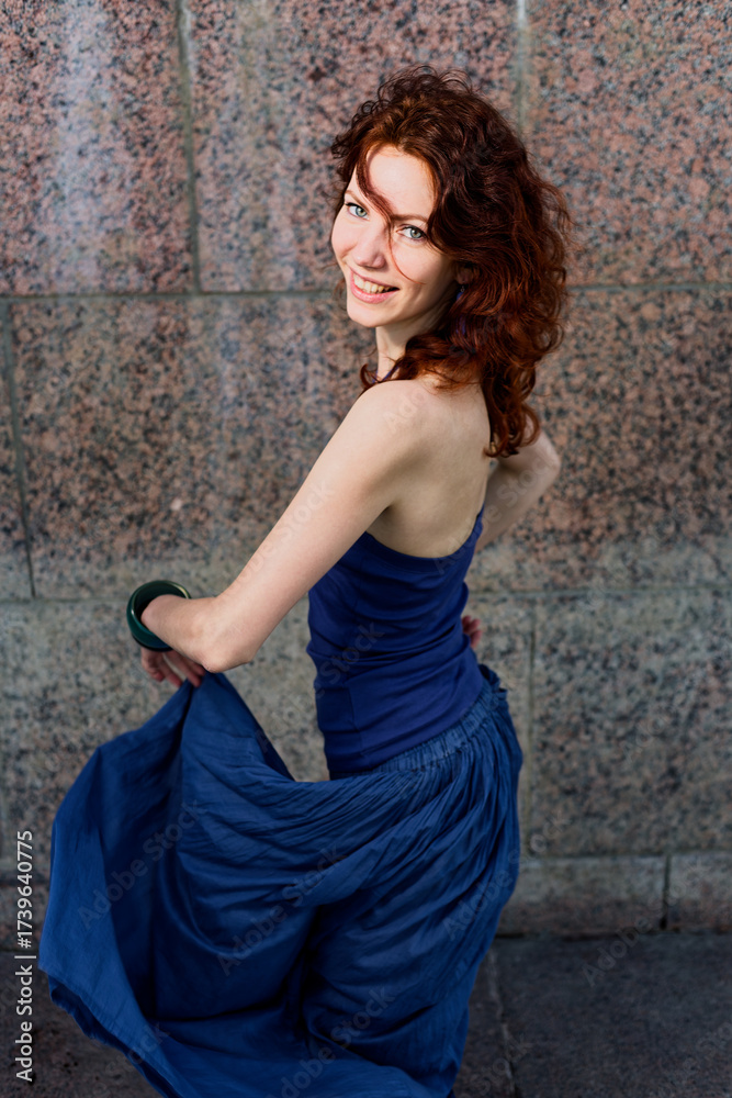 Fototapeta premium Smiling Woman with Red Hair Twirls Blue Skirt Against Granite Wall