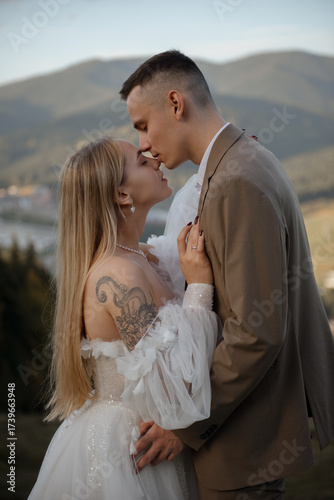 A beautiful, happy young couple in love on a wedding walk and photo shoot high in the mountains. The groom in a jacket and suit and the bride in a white dress at a resort. Kissing and hugging