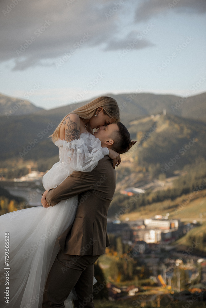 Fototapeta premium A beautiful, happy young couple in love on a wedding walk and photo shoot high in the mountains. The groom in a jacket and suit and the bride in a white dress at a resort. Kissing and hugging