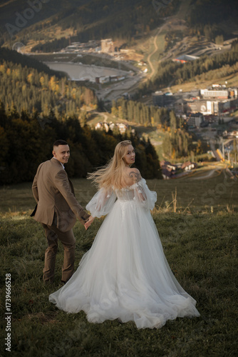 A beautiful, happy young couple in love on a wedding walk and photo shoot high in the mountains. The groom in a jacket and suit and the bride in a white dress at a resort. Kissing and hugging