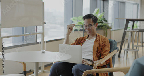 Senior Asian businessman wearing headset celebrates success during online meeting in modern co-working space. Remote work and business communication concept	