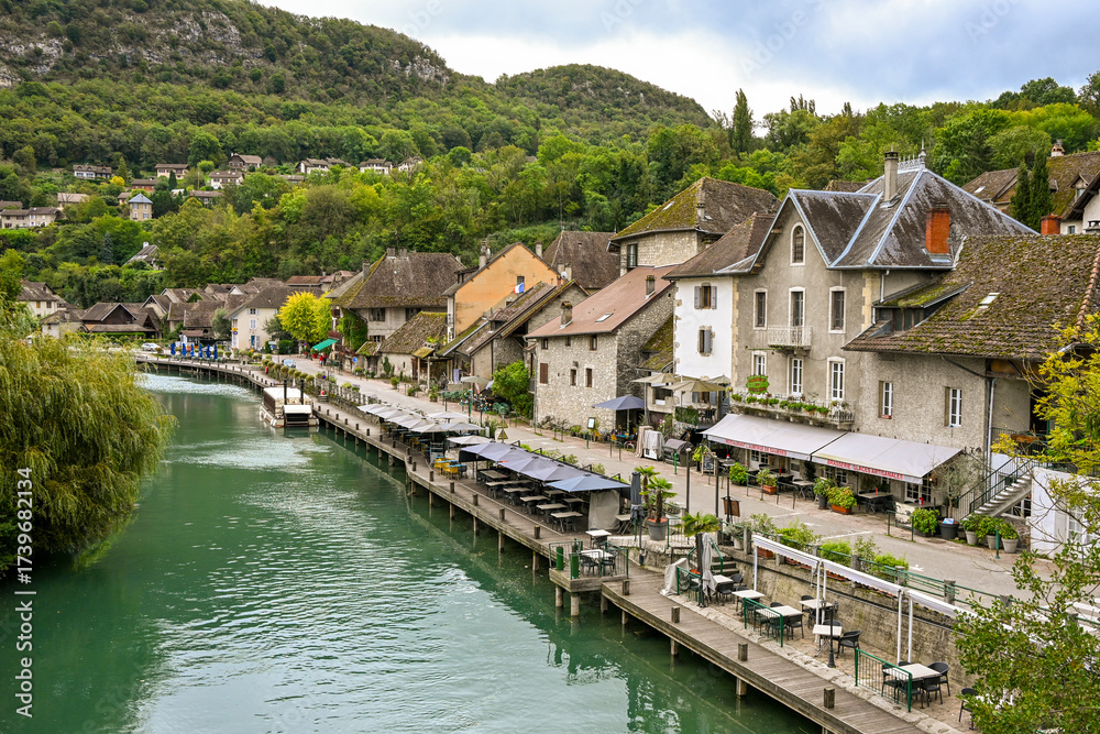 Fototapeta premium A charming riverside view of Chanaz, France, with traditional houses, waterfront cafés, and lush green hills in the background.