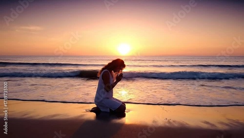 Woman kneeling on beach at sunset and praying by the ocean  