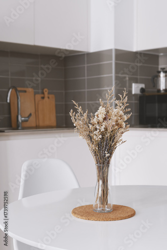 Modern minimalist kitchen with a glass vase of dried flowers on a round white table. Cozy neutral home interior with natural light, soft tones, and contemporary design.