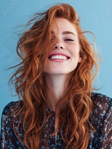 A cheerful red-haired woman with a vibrant smile poses against a bright blue background, exuding happiness and confidence.
