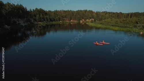 Aerial view of two young girls in life jackets paddling down river on SUP boards. Magnificent summer landscape with pine forest against blue sky. Concept of silence and pacification