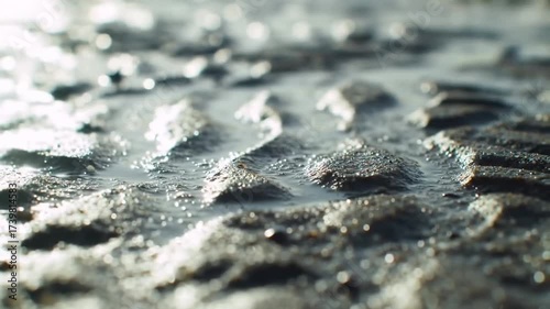 Shallow, sunlit water flows over a textured, dark sand surface, creating ripples and reflections. The close-up perspective emphasizes the detailed texture and the interplay of light and water