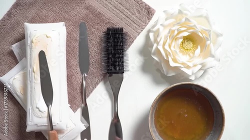 Flatlay showcasing spa items  towels,  rectangular objects with creamy substance, a bowl of liquid, a brush, and an artificial flower