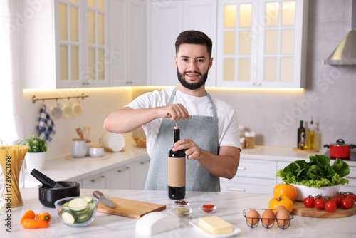Photos Smiling man opening bottle of wine while cooking at table in kitchen