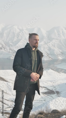 Vertical video. Attractive young man with beard and hair in a bun walks along a winter mountain seashore with snowy peaks behind. Handsome guy dressed in white sneakers and a jacket.