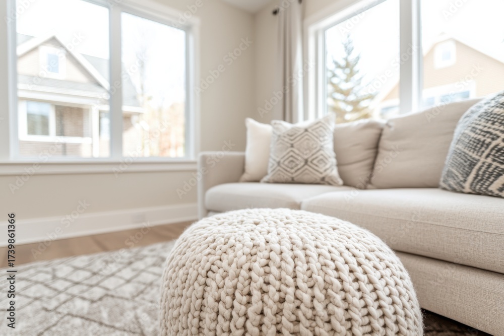 Fototapeta premium A cozy living room featuring a light-colored sofa, decorative pillows, and a knitted pouf, bathed in natural light from large windows.