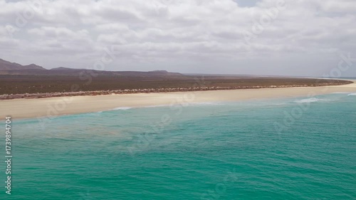 Aerial footage of Santa Monica Beach,showcasing endless white sand, turquoise waves, and a pristine,background volcanic mountain and desert,Boa Vista, Cape Verde.