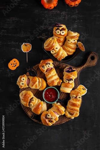 Halloween food. Scary sausage mummies in dough with funny eyes on dark background. Funny decoration. Top view