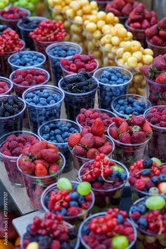 An inviting display of various fresh berries, including blueberries, raspberries, strawberries, and mulberries, portioned in plastic cups at a market.