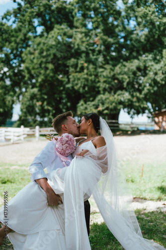 Bride and groom kissing outdoors in wedding attire. The groom is holding the bride in his arms.