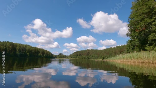 Point of view shot gliding forward at water level across a calm summer lake with ripples, reeds, forest reflections and white clouds in the blue sky
