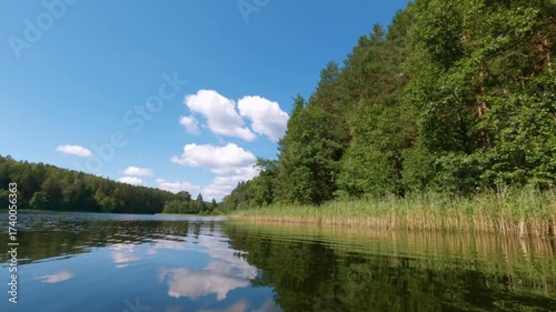 Point of view shot gliding forward at water level across a calm summer lake with ripples, reeds, forest reflections and white clouds in the blue sky