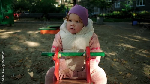 Child plays on a red playground toy in a sunny park surrounded by trees and benches