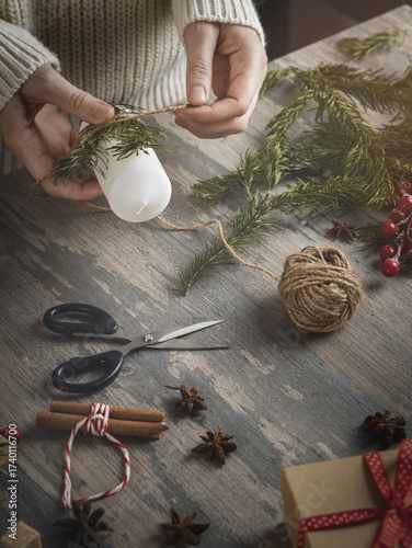 Female hands making Christmas decoration with candle, spruce needles, jute twine on a wooden background, vertical image.
